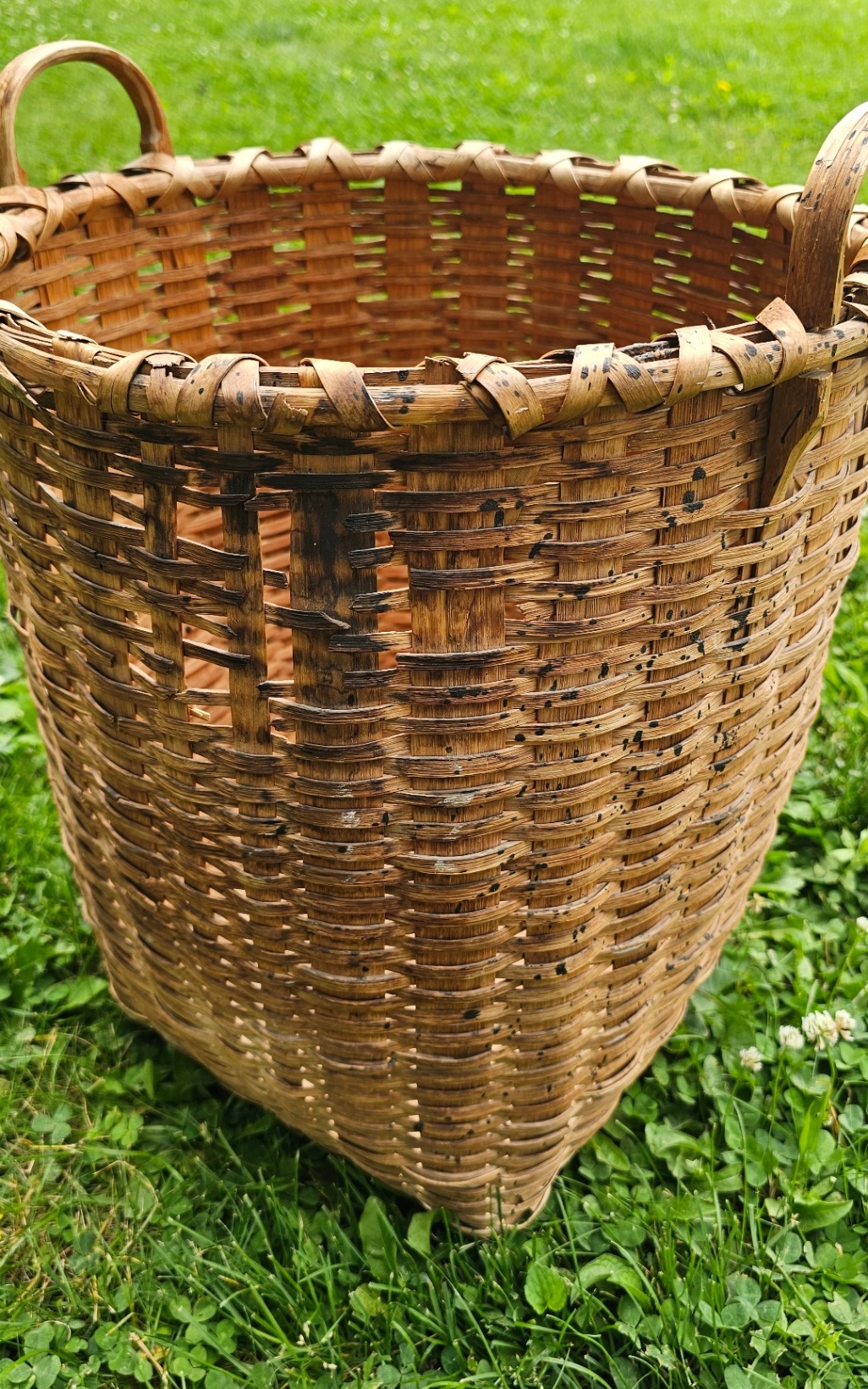 A large Taghkanic ash splint basket with two handles, sides darkened and stained from a house fire but still structurally intact, sitting on grass.