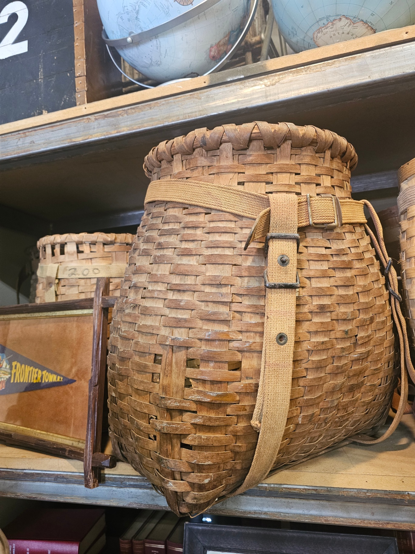 A pot-belly pack basket with original canvas straps on a shelf at a Hudson antique shop, with other ash baskets visible nearby.